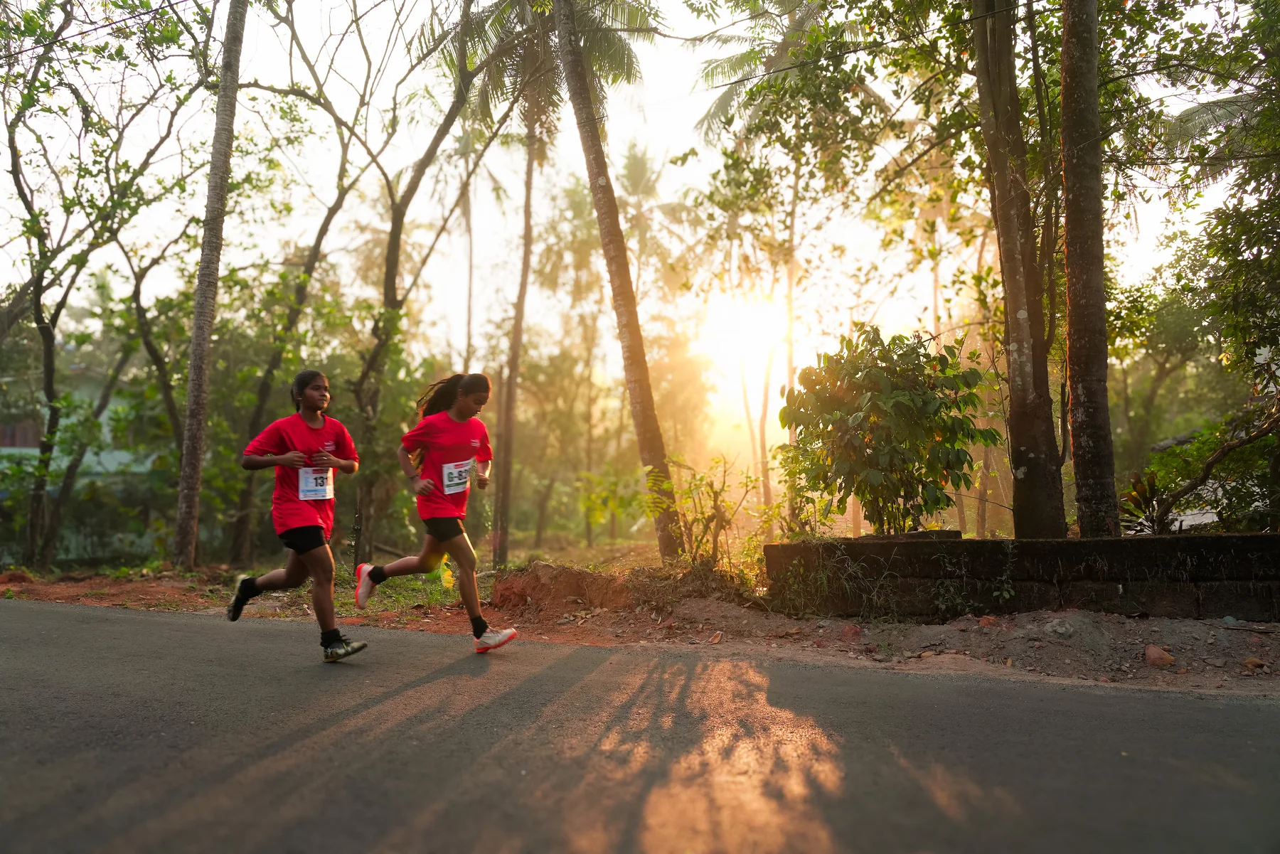 Two runners crossing a sunlit road through Peravoor